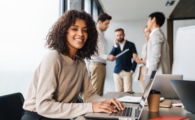 Attraktive afrikanische junge selbstbewusste Geschäftsfrau sitzt am Bürotisch mit einer Gruppe von Kollegen im Hintergrund, arbeitet am Laptop-Computer